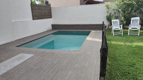 Une piscine rectangulaire avec une terrasse carrelée, entourée d’un mur blanc et d’une clôture en bois. On aperçoit deux chaises blanches à l’arrière-plan.