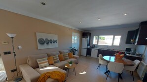 Een moderne woonkamer met een bank, salontafel en een eethoek met tafel en stoelen. In de achtergrond is een keuken met een koelkast en magnetron zichtbaar.