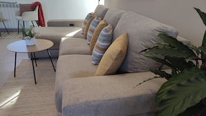 Een woonkamer met een beige bank, kussens met strepen en effen kleuren, een klein rond tafeltje met een plant, en een grote groene plant op de voorgrond.