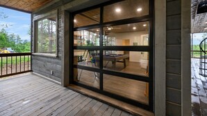 A covered outdoor area with a pool table, wooden deck, and large windows overlooking a wooded area. - 5 Bedroom Sleeps 24 , Pets OK, Hot Tub, Game Room. (Broken Bow)