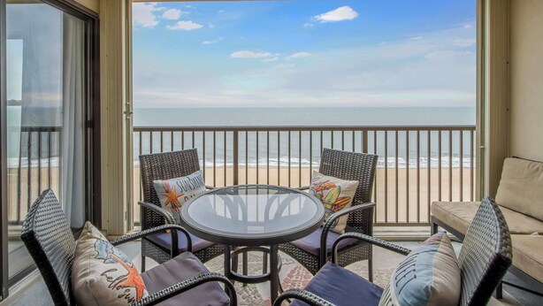 A balcony with wicker furniture, a glass table, and a view of the beach. - Sandpiper Dunes 1009 (Ocean City)