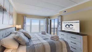 A hotel room with a large bed, a TV, a balcony with curtains, and a sign for Ocean City Vacation Homes. - Sandpiper Dunes 1009 (Ocean City)