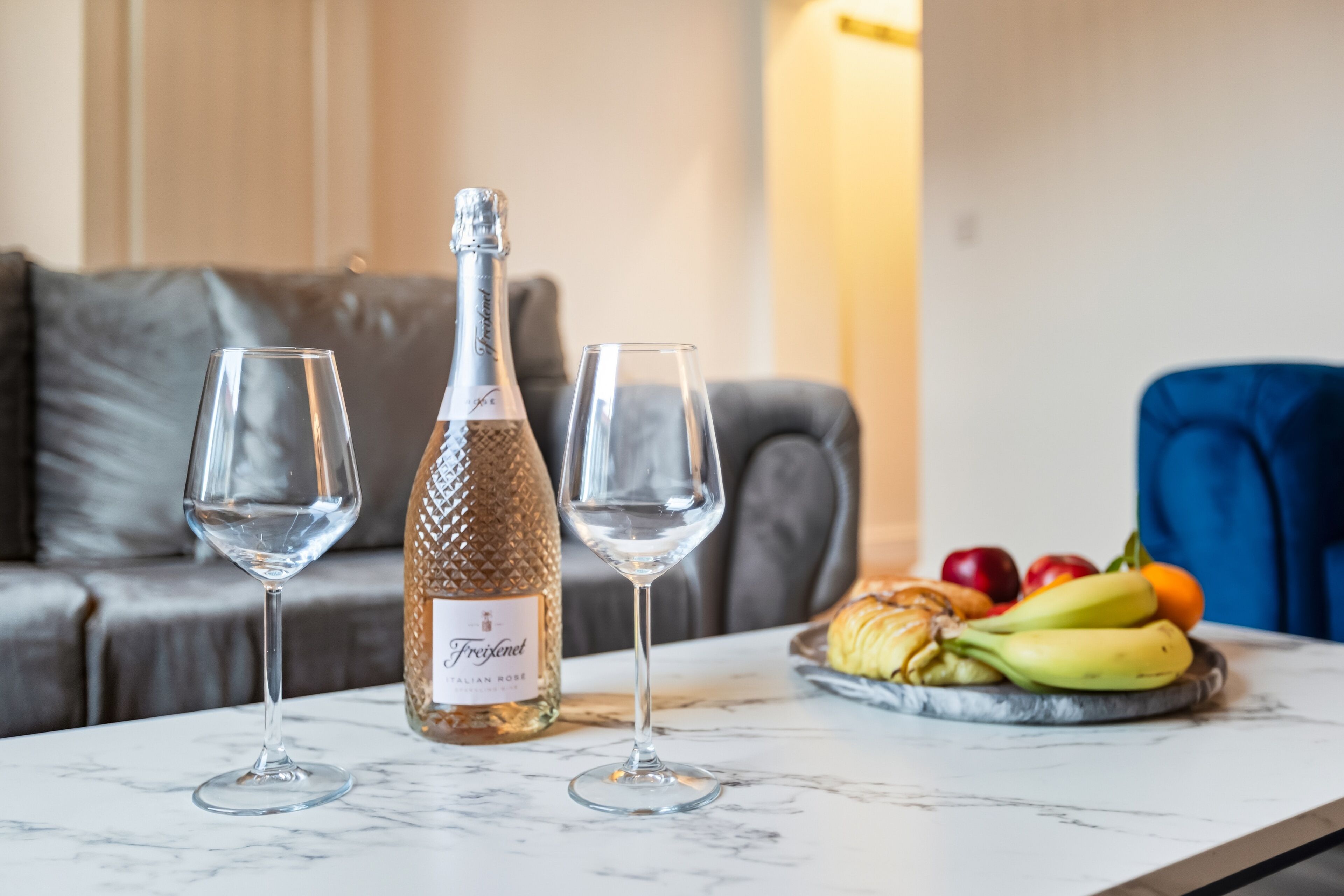 Uma mesa de mármore com duas taças de vinho, uma garrafa de vinho rosé italiano Frejolent e um prato de frutas.