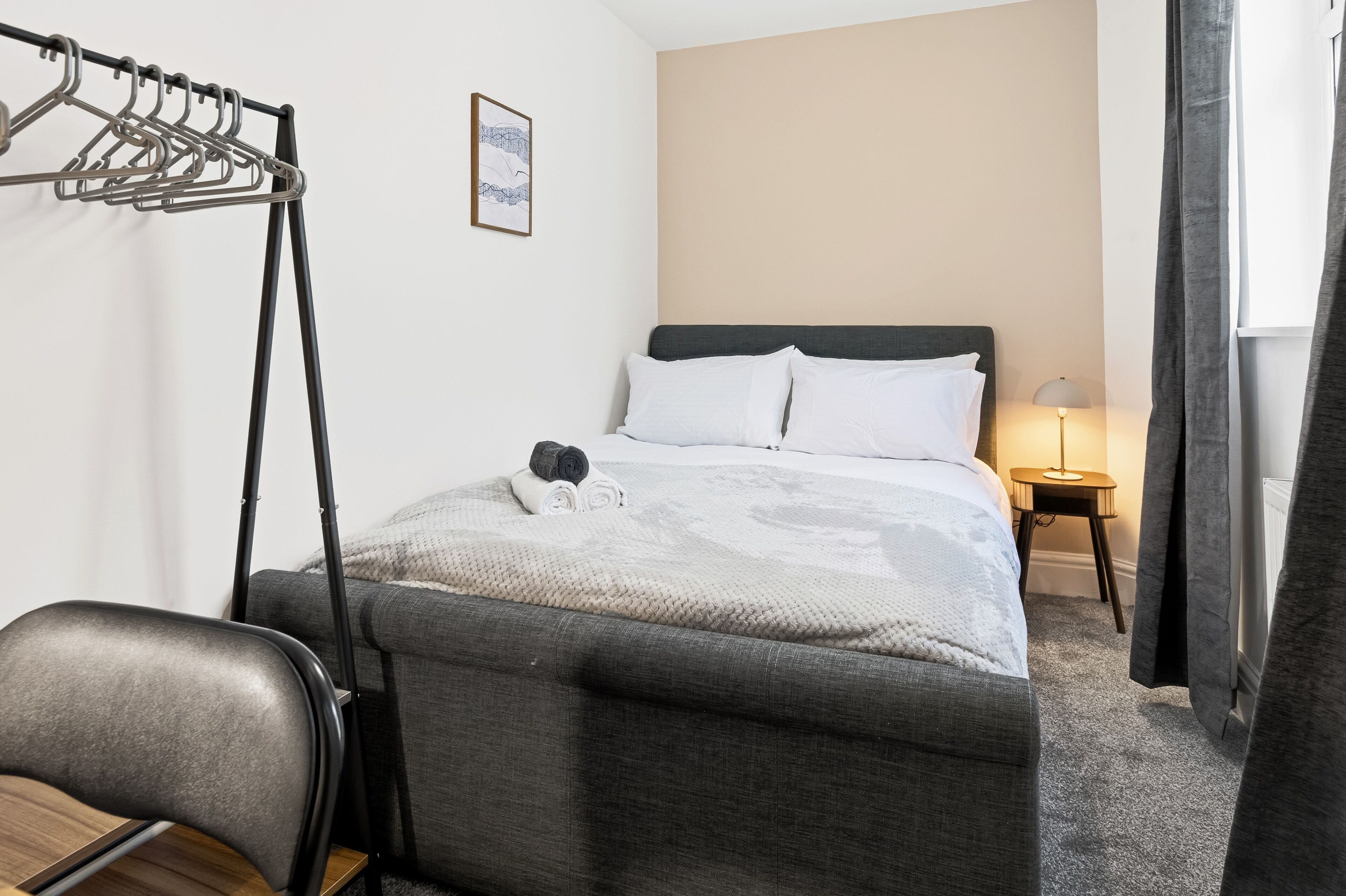 A hotel room with a bed, a chair, a coat rack, a small table with a lamp, and a picture on the wall.