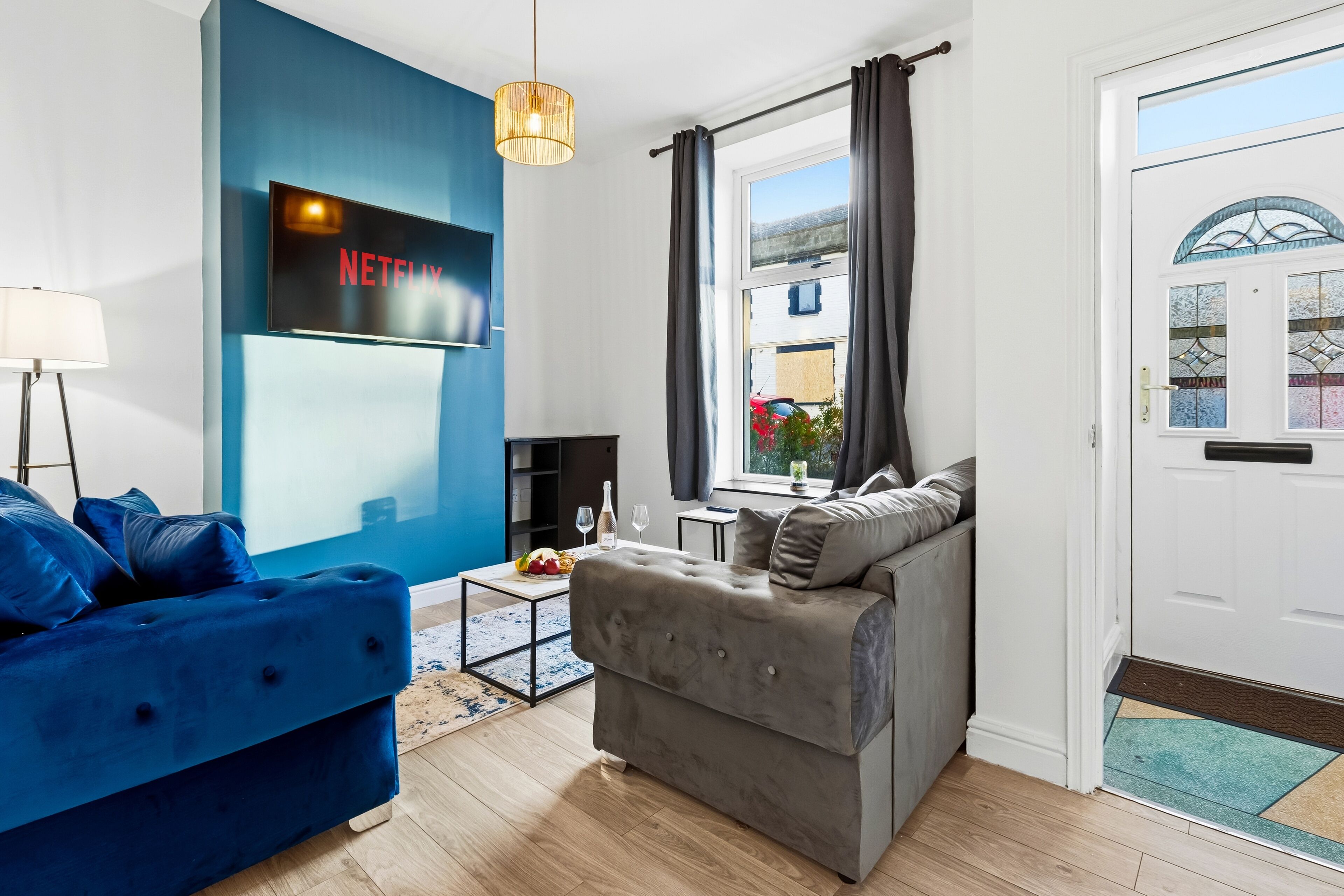 Een moderne woonkamer met een blauwe bank, een grijze bank, een flatscreen televisie bevestigd aan een blauwe muur, een bijzettafeltje met een schaal fruit, en een raam met gordijnen.
