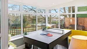 Una mesa de ping pong en una habitación con ventanas amplias, un sofá amarillo y una lámpara.