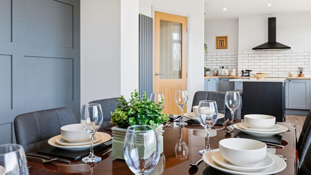 Un comedor moderno con una mesa de madera dispuesta para seis comensales, rodeada de sillas tapizadas en gris. Una planta en maceta aporta un toque de verdor.
