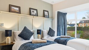 Habitación de hotel con dos camas, una mesita de noche con lámparas, una estantería con fotografías enmarcadas y vistas al exterior.