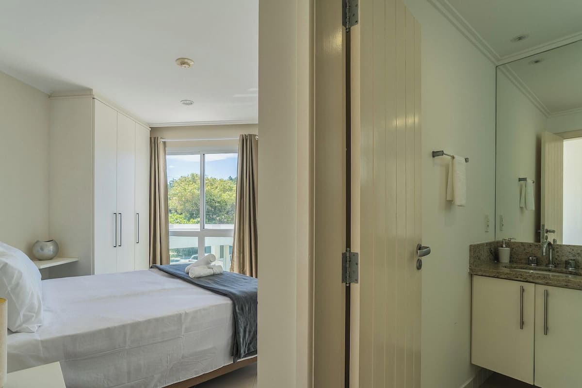 A bedroom with a bed, a bedside table, a window with curtains, and a view of greenery outside.