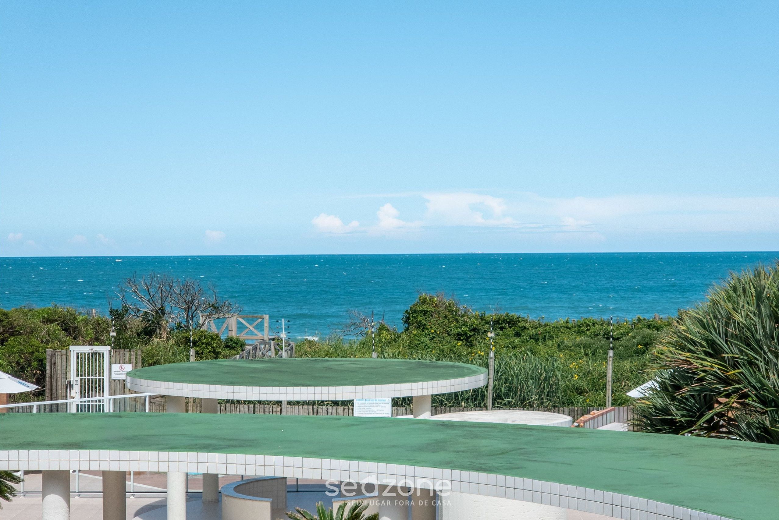 A view of a coastal area with a circular green space, a wooden fence, and a clear blue sky.