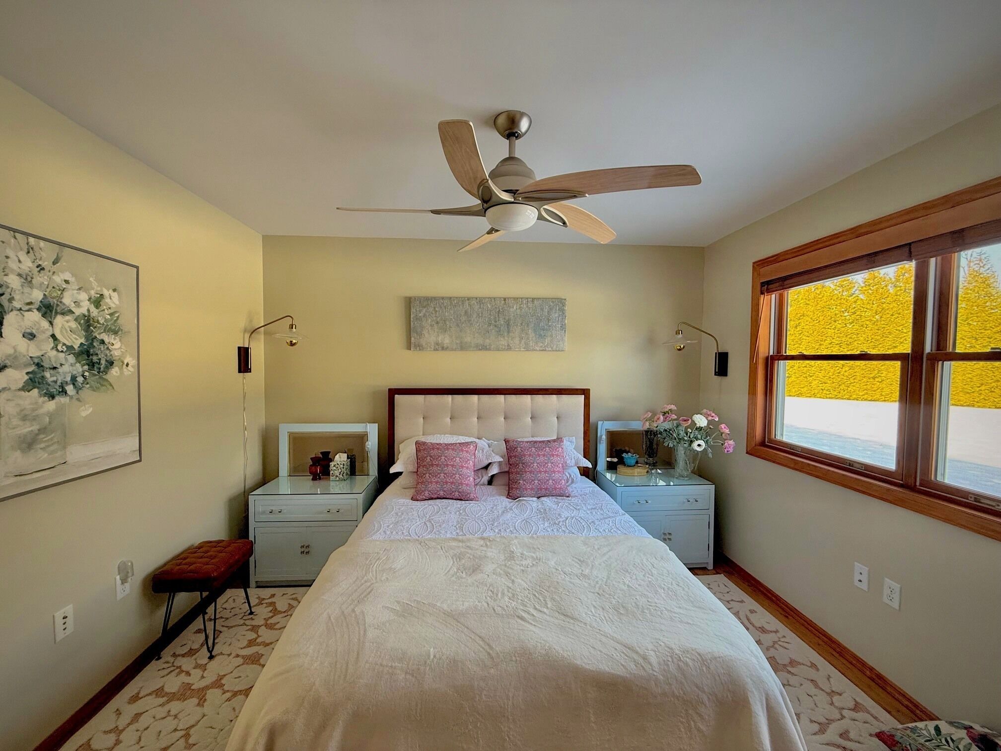 A bedroom with a bed, a ceiling fan, bedside tables, a nightstand, a window, and a painting on the wall.