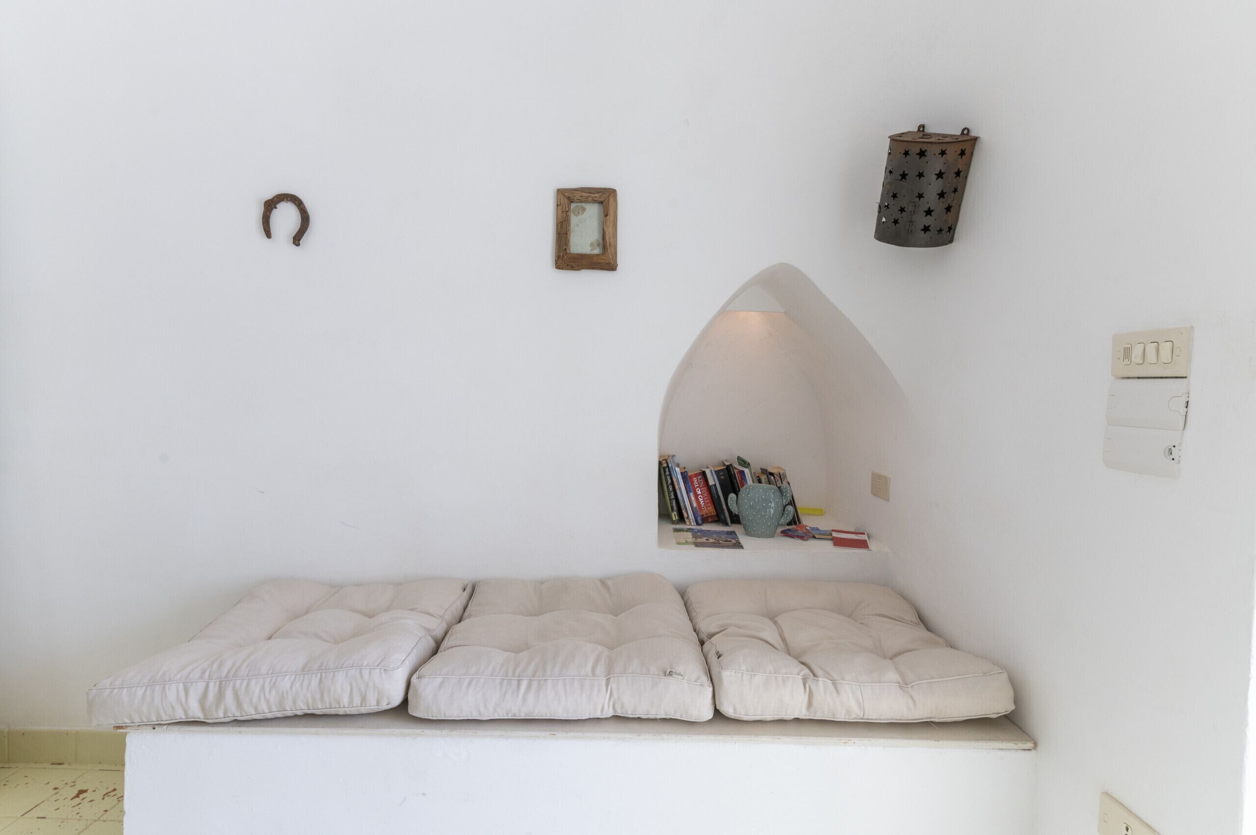 A minimalist room with a cushioned bench, a small framed picture, a star-patterned lantern, and a shelf with books and a mug.