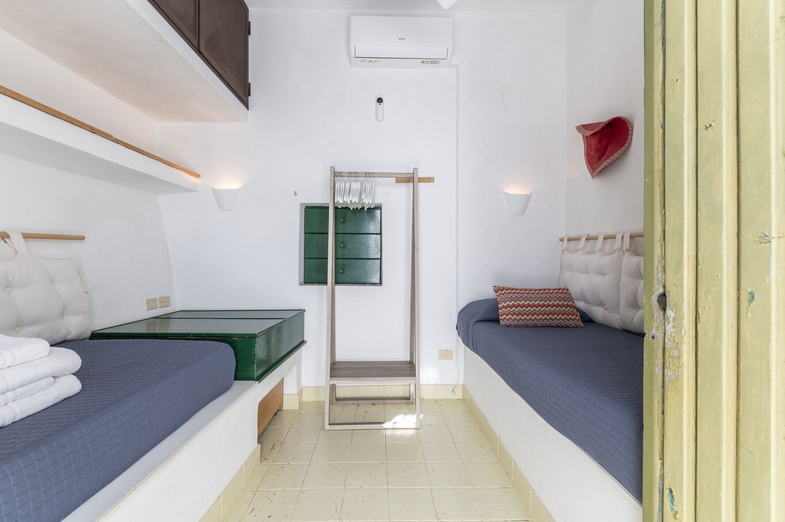 Een kleine, compacte kamer met twee bedden, een groene dressoir, een spiegel en een wandairconditioner.