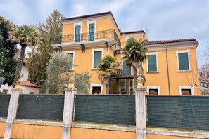 A two-story building with a balcony, surrounded by a green fence and palm trees. - Beach holiday at Venice Lido: comfort and charm in Art Nouveau style (Venezia)