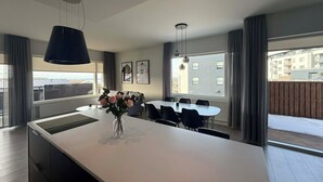 Cozinha moderna com ilha central, área de jantar com cadeiras, luminárias pendentes e grandes janelas com cortinas.