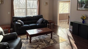 Ein Wohnzimmer mit einem dunkelbraunen Ledersofa, einem Holztisch und einem gemusterten Teppich. Ein Fenster mit Vorhängen, eine Tür zu einem anderen Raum und ein Bild an der Wand sind ebenfalls zu sehen.
