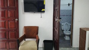 A room with a wooden door, a television mounted on the wall, a chair, a trash bin, and a view of a toilet through an open door. - Kom Shot 38 (Cancún)