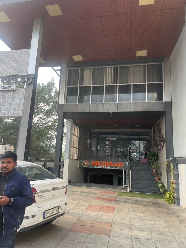 Entrada de un edificio con un cartel que dice 'Mezbaan Banquet & Restaurant' y un coche blanco estacionado al frente.