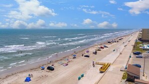 A beach with a long stretch of sand, umbrellas, and beach chairs. There are buildings and a parking lot adjacent to the beach. - Cozy Beach Retreat Near Whitecap & Padre Island (Corpus Christi)