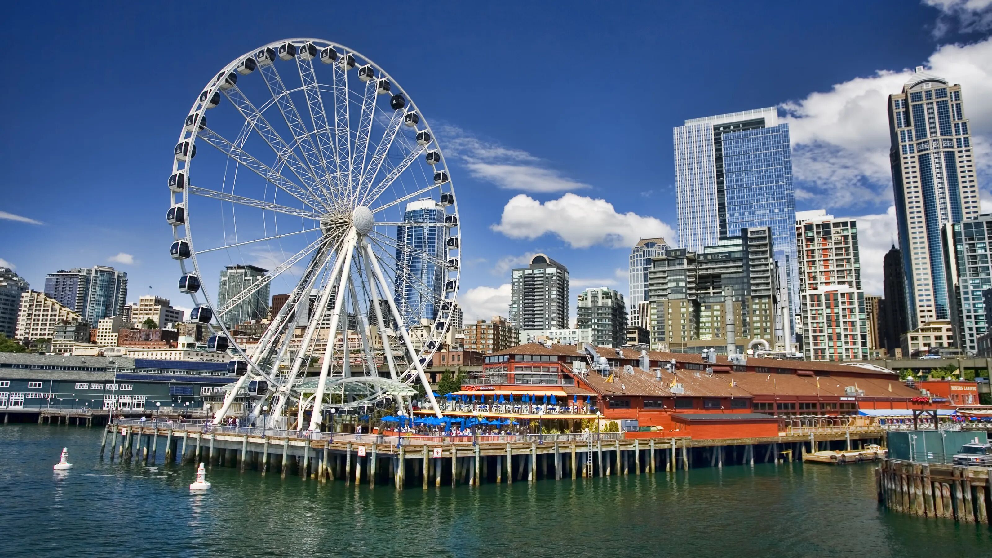 Paisagem urbana à beira-mar com uma roda-gigante em destaque, edifícios modernos altos e um píer com restaurante.
