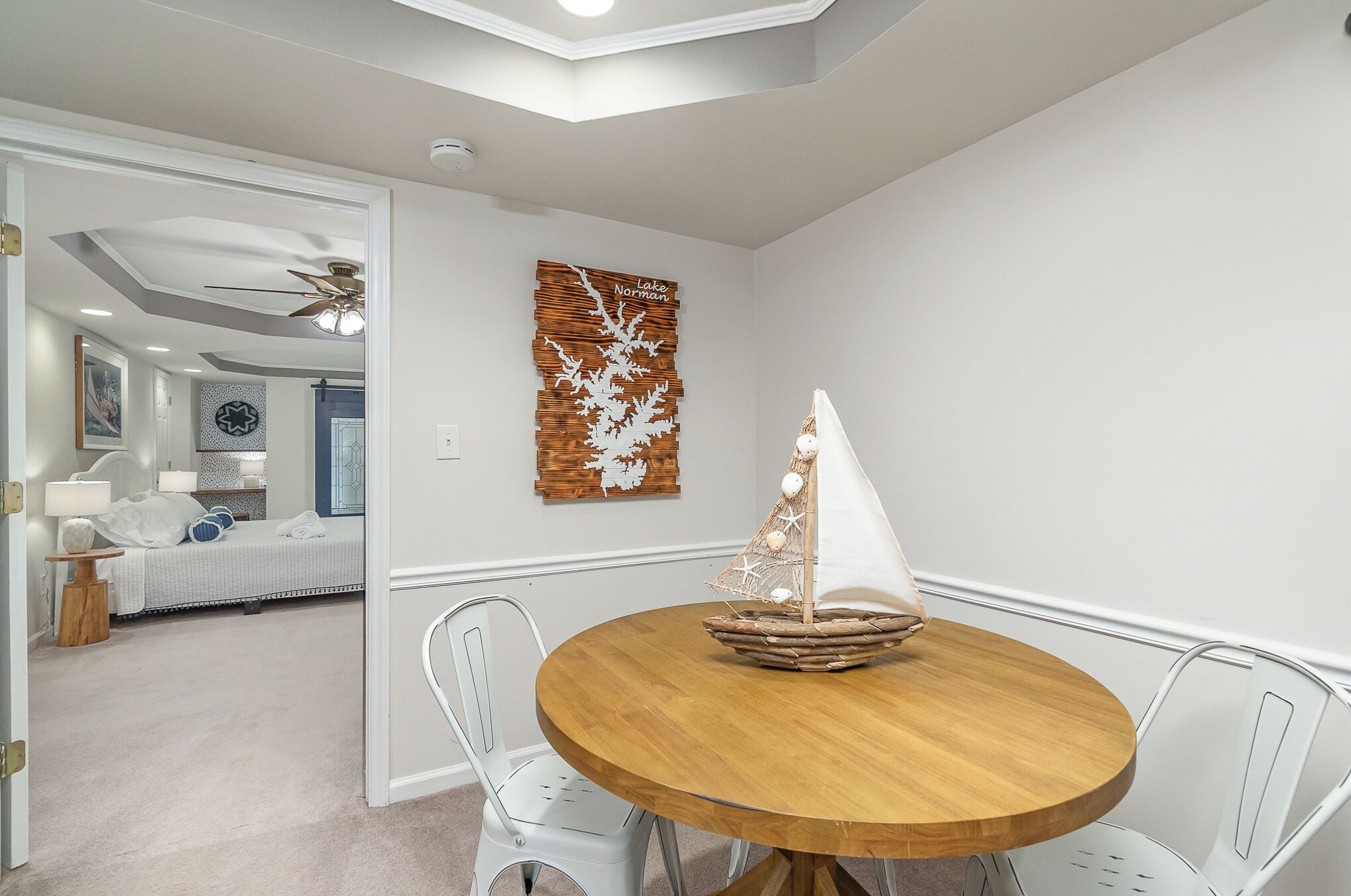 A dining area with a round wooden table and white metal chairs. A decorative sailboat is placed on the table. The room has white walls and a ceiling fan.