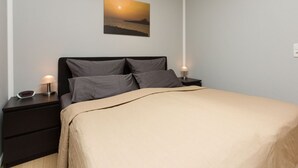 Een opgemaakt bed met een beige deken, twee nachtkastjes met lampen en een aan de muur hangend schilderij van een zonsondergang boven water.