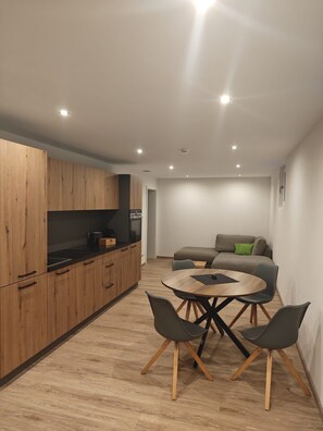 A modern kitchen with wooden cabinets, a dining area with a round table and chairs, and a sofa in the background. - 75-square-meter apartment for an installer (Garching an der Alz)