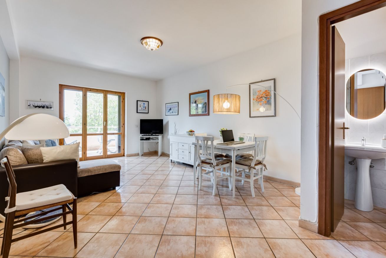 A spacious living area with a sofa, dining table, and chairs, a kitchen in the background, and a large window.