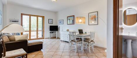 A spacious living area with a sofa, dining table, and chairs, a kitchen in the background, and a large window.
