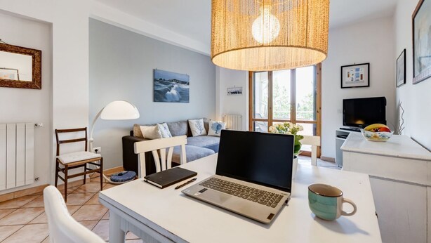 Un salón moderno con un ordenador portátil sobre una mesa, un sofá, una silla, una lámpara y un ventanal grande.