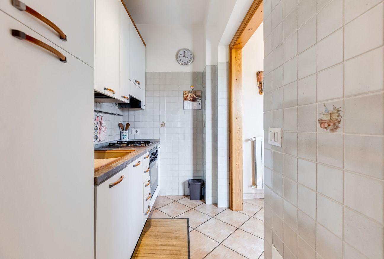 Une cuisine étroite avec des meubles blancs, un fourneau et une horloge au mur.