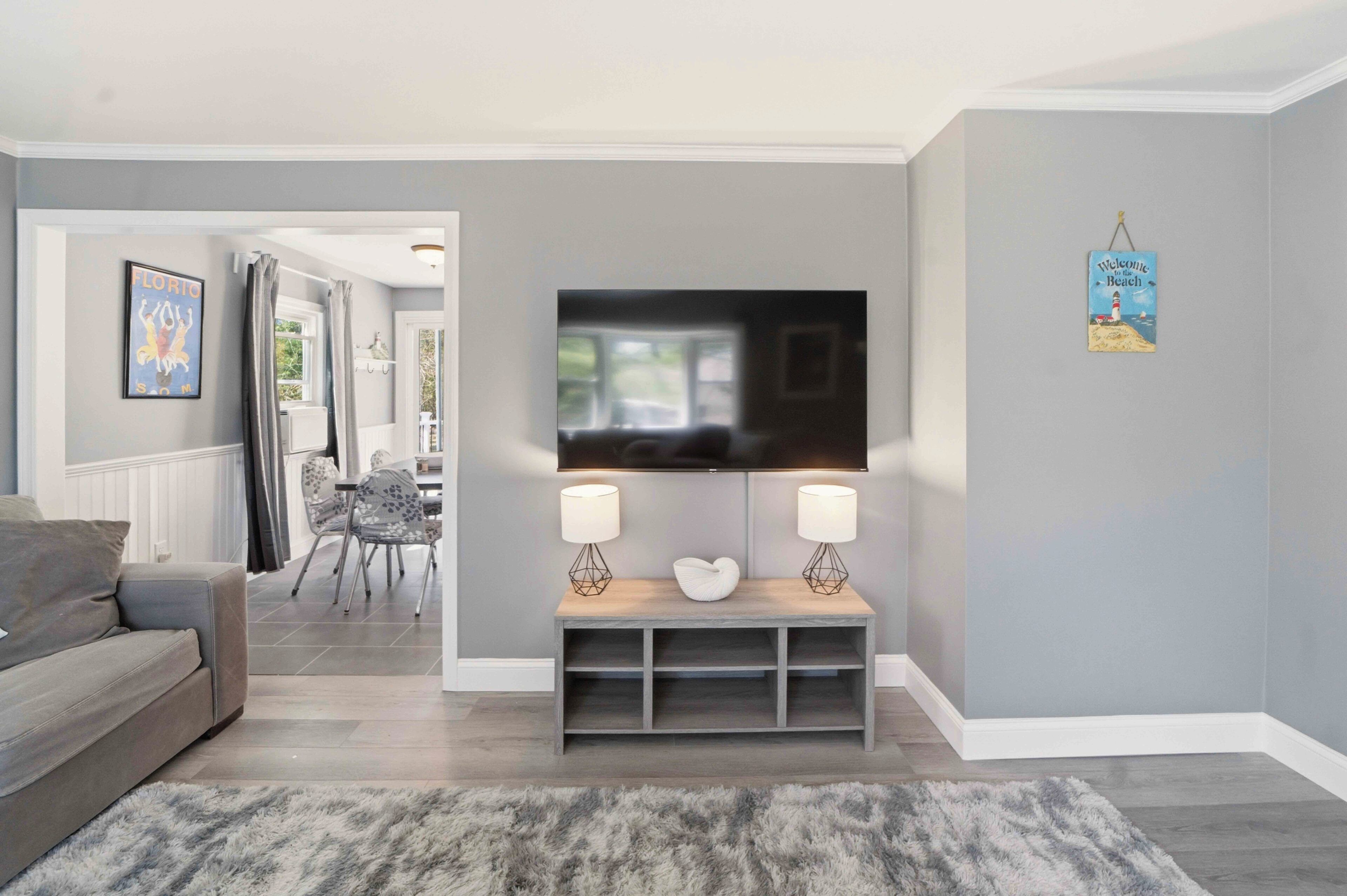 A living room with a grey sofa, a TV on a stand, a dining area with chairs, and a grey rug.