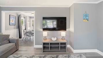 A living room with a grey sofa, a TV on a stand, a dining area with chairs, and a grey rug.