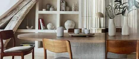 A dining area with a round table, two wooden chairs with cushions, a bookshelf with various items, and a decorative plant.