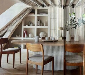 A dining area with a round table, two wooden chairs with cushions, a bookshelf with various items, and a decorative plant. - Luxurious Apartment Place Vendome - Rue de la Paix (Paris)