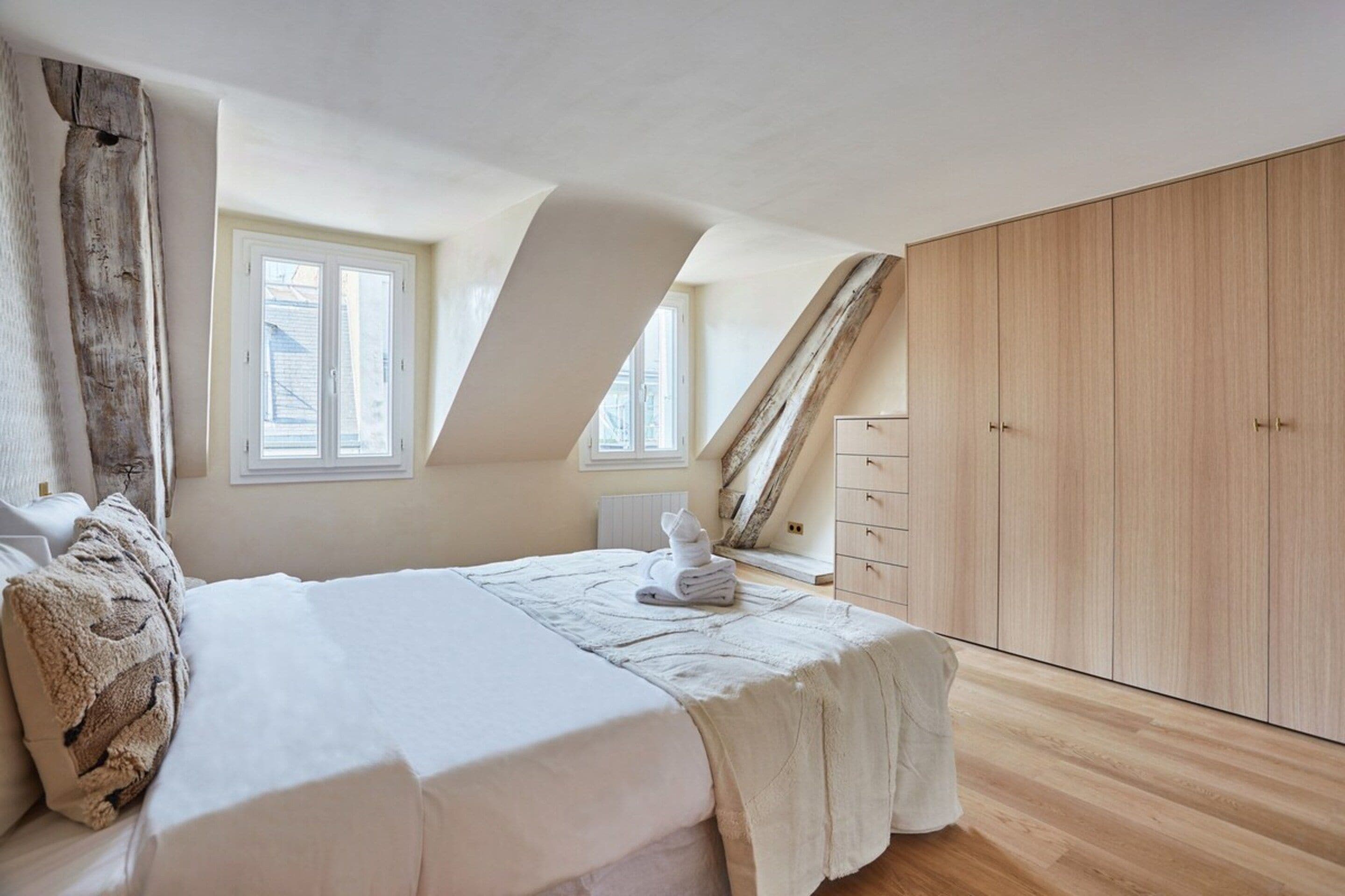 A bedroom with a sloped ceiling, a large bed, a wooden wardrobe, and a window.