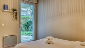 Ett hotellrum med en säng, ett fönster, en väggmonterad luftkonditioneringsanläggning, en taklampa och en liten hylla med en växt och en låda märkt "PARIS".