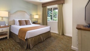 Habitación de hotel con una cama, dos mesitas de noche con lámparas, un ventilador de techo, una ventana con cortinas y un televisor sobre un mueble de madera.