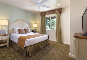Ein Hotelzimmer mit einem Bett, zwei Nachttischen mit Lampen, einem Deckenventilator, einem Fenster mit Vorhängen und einem Fernseher auf einem Holzkasten.