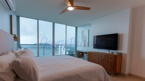 A modern bedroom with a large bed, a desk, a television, and a view of the city through large windows. - 23M Spectacular New Penthouse Oceanview (Panama City)