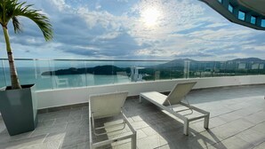 A balcony with two white wicker chairs, a potted palm tree, and a glass railing overlooking a scenic view of a body of water and distant mountains. - 23M Spectacular New Penthouse Oceanview (Panama City)