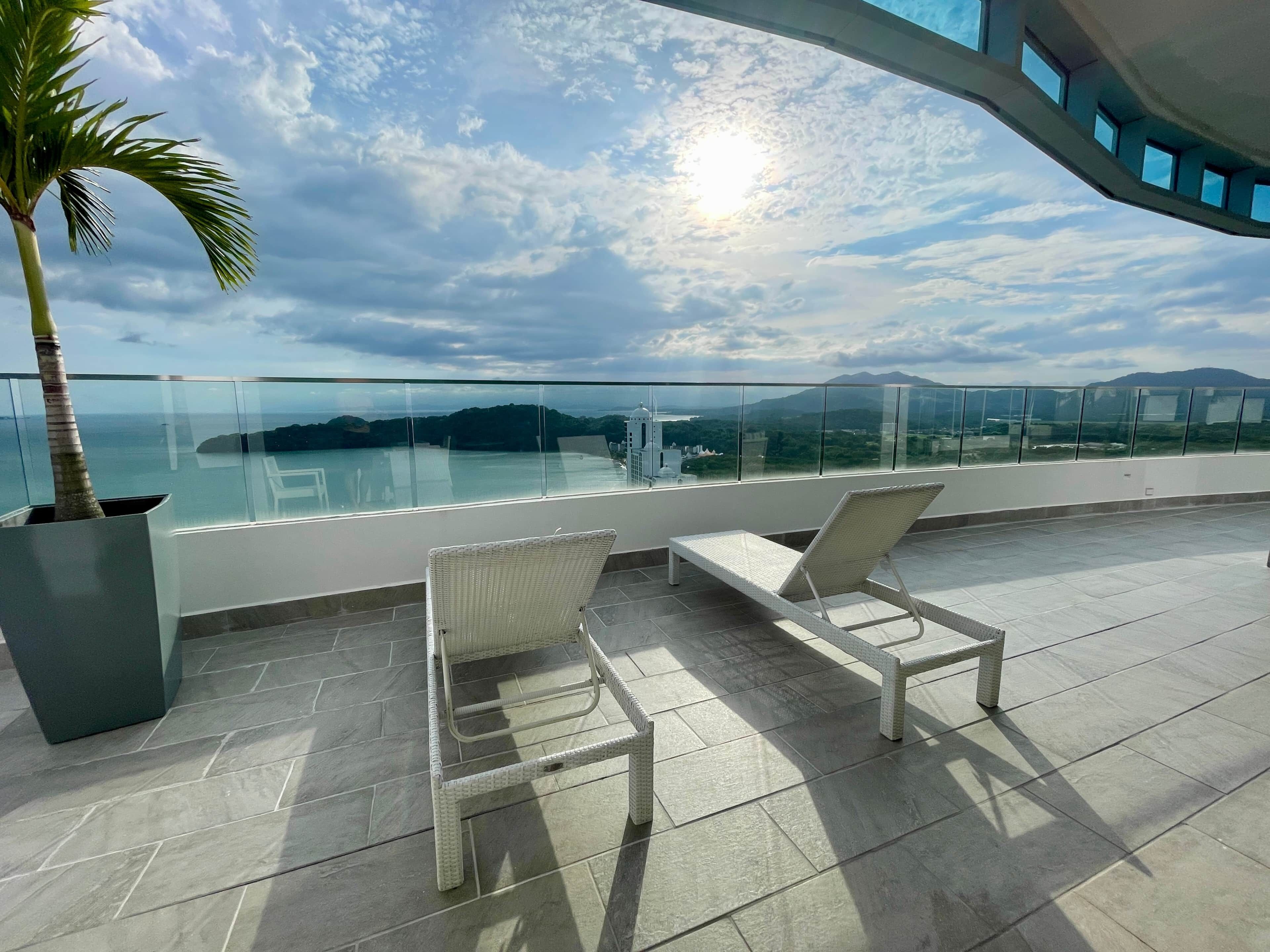 Un balcón con dos sillones de mimbre blanco, una palmera en maceta y barandilla de cristal con vistas panorámicas a un cuerpo de agua y montañas distantes.
