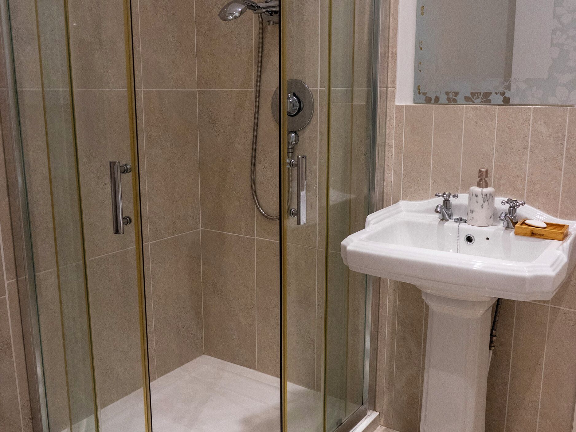A bathroom with a glass-enclosed shower, a white sink with two faucets, and a soap dispenser.