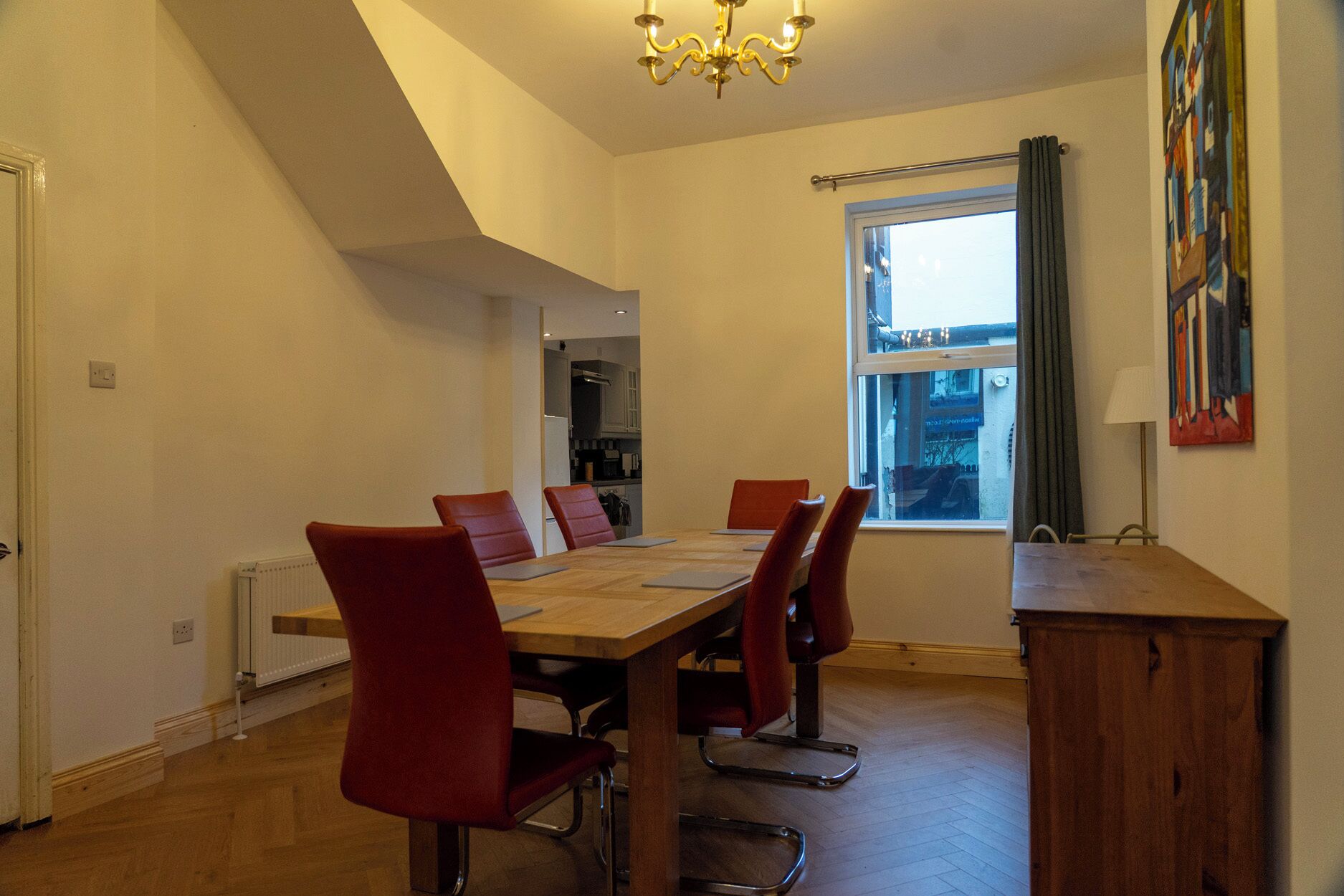 A dining area with a wooden table and red chairs, a chandelier, a window with curtains, and a painting on the wall.