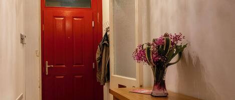 A hallway with a red door, a wooden bench, a small table with a vase of flowers, and a coat hanging on the wall.