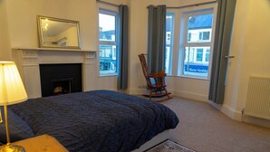 A bedroom with a bed, a rocking chair, a fireplace, and large windows. - The Red Door (Bangor)