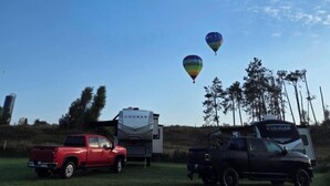 Un cielo azul despejado con cuatro globos aerostáticos y una autocaravana grande en un campo cubierto de hierba.