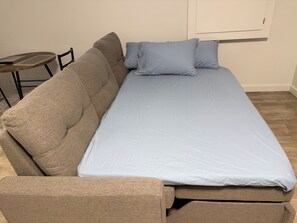 Ein Einzelbett mit hellblauer Matratze und Kissen, ein beiges Sofa und ein kleiner Holztisch.