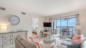 A spacious living room with a large window, a flat-screen TV, and a balcony with a table and chairs. - Carlos Pointe #624 (Fort Myers Beach)