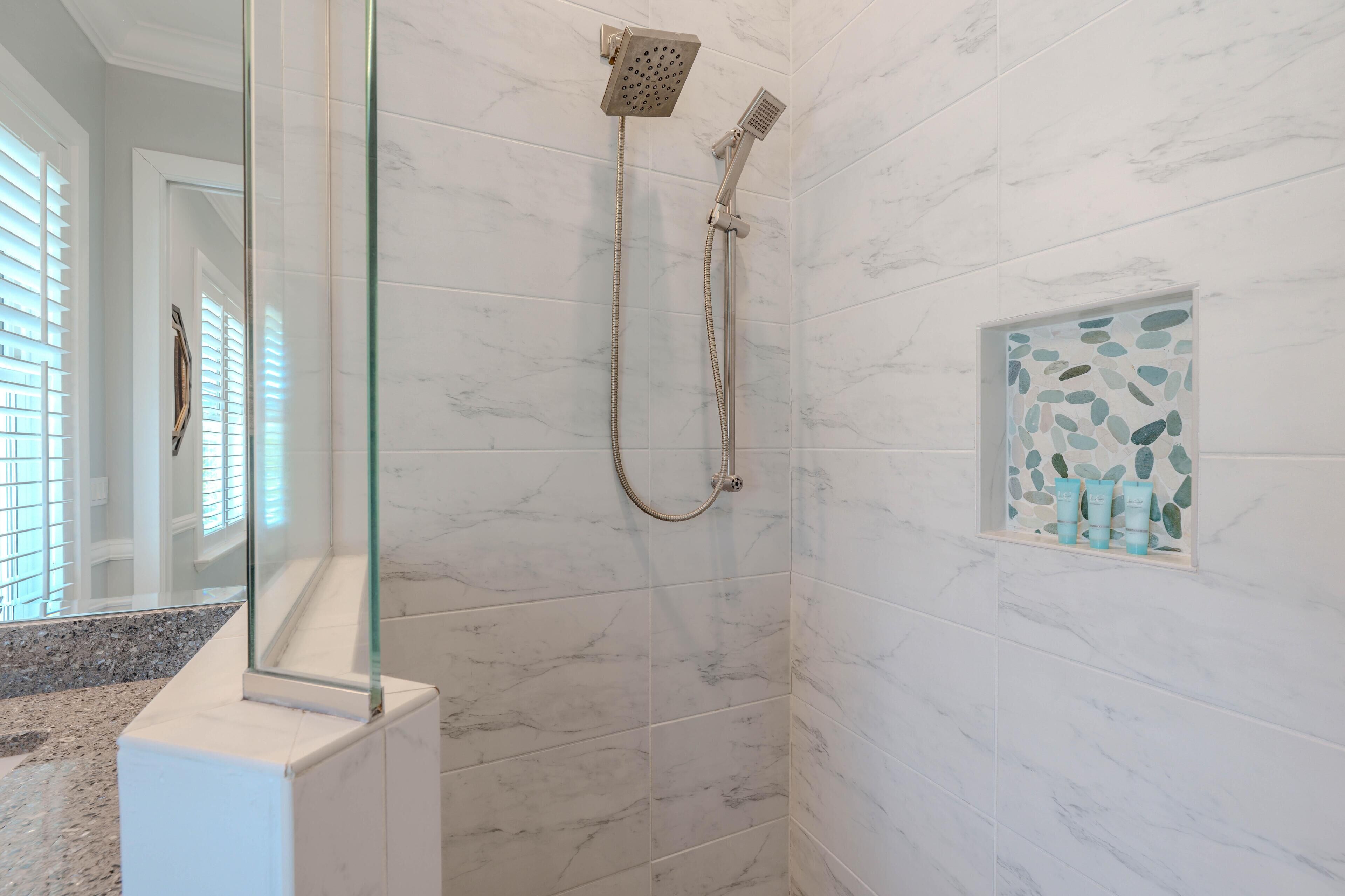 A bathroom with a glass shower enclosure, white marble tiles, and a small shelf with toiletries.